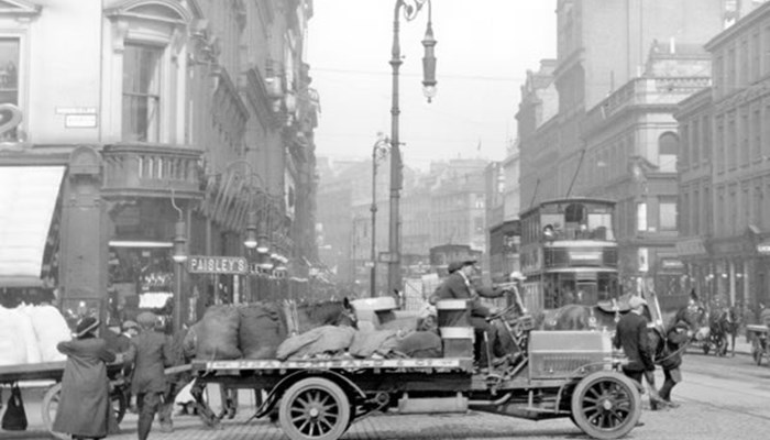 A black and white photo of a horse and carriage being loaded, with trams and people in the background along with tall tenement shops.