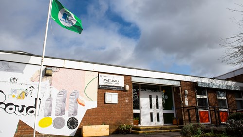 exterior shot of a community centre made of brown brick. there is a sign that says castlemilk community centre in white and black. there is a green flag on a flagpole outside the venue. there is a mural on the side of the build with the work proud printed beside the entrance