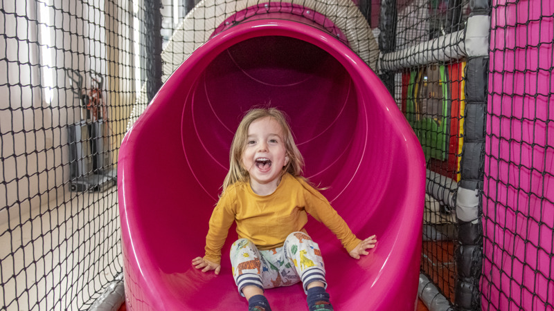 A young child laughing and smiling as they come out of the end of a dark pink tunnel slide.