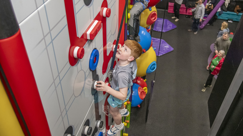 A young boy with red hair looking up as he climbs a multi-coloured climbing wall.
