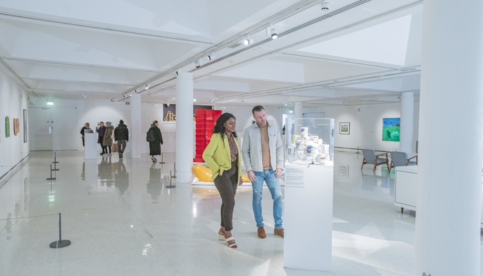 Two visitors in central galleries at GoMA look at an object in a glass display case. The gallery is a white cube with a diverse offer of objects on display including paintings and sculptures in the background