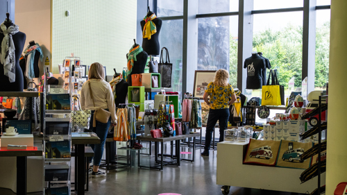 A store interior with various items on display, including bags, scarves, books, and other merchandise. Two people are browsing the items. The store has large windows allowing natural light to enter.