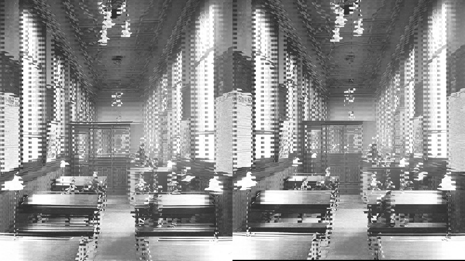 A black and white photograph of inside Bridgeton Library. There are three rows of tables with lamps, with 8 children reading and a librarian at a curved wooden desk.