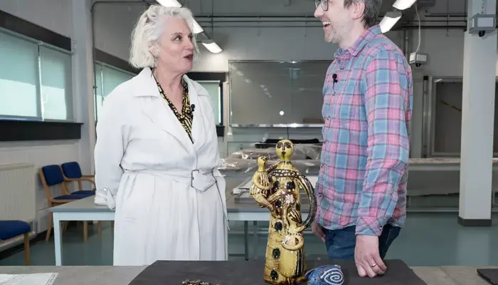 Two people stand in a bright studio-like space with exposed lighting overhead, each wearing a small microphone clipped to their clothing. The woman on the left is dressed in a white coat, and the man on the right wears a plaid shirt and jeans. In front of them, a table displays several art and artifact pieces, including a gold-toned ceramic sculpture with intricate figures, a blue spiral-patterned animal-shaped object, a beaded necklace, and a large protected drawing of a curled cat. 