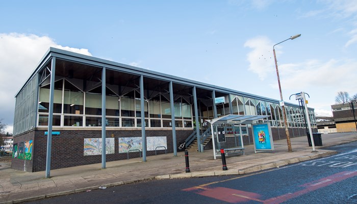 Exterior of Pollokshaws Library which is large, rectangular, flat roofed 1960s building. There a 7 slim iron pillars near the entrance. There is a bus stop directly outside. The photograph also shows a sunny, blue sky day.