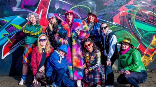 A group of nine women, dressed in brightly coloured clothes, pose in front of a graffiti backdrop. They are collectively known as Scottish hip hop group Female Funk Fruition