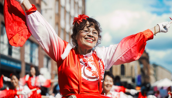 A smiling woman wearing glasses and a vibrant red and white traditional outfit dances joyfully in the street with her arms raised, holding a red cloth and performing alongside other dancers.