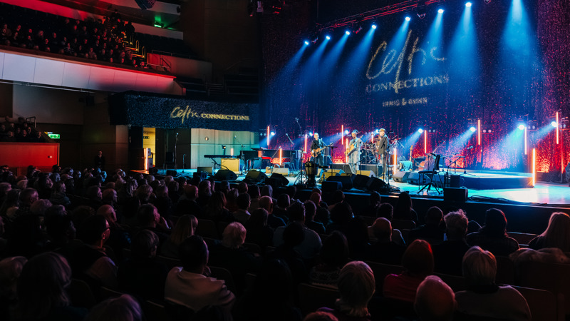 Audience seated in a concert hall watching a live band perform on a brightly lit stage under blue and red spotlights.