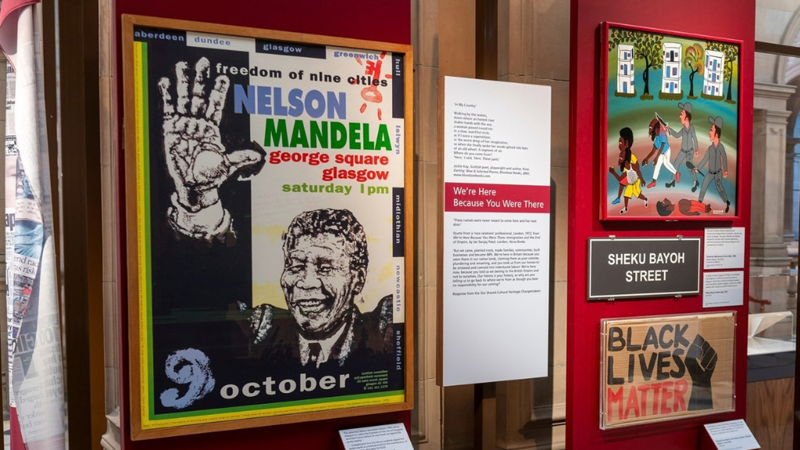 A museum display featuring a poster of Nelson Mandela promoting a “Freedom of Nine Cities” event in Glasgow, alongside an illustrated artwork, a “Sheku Bayoh Street” sign, and a “Black Lives Matter” sign.