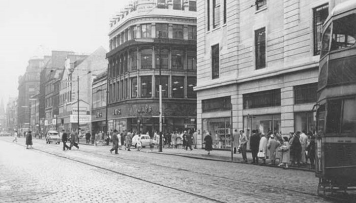 A busy Glasgow street, with lots of people walking around, cars and buses/trams on the street, with large shop buildings.