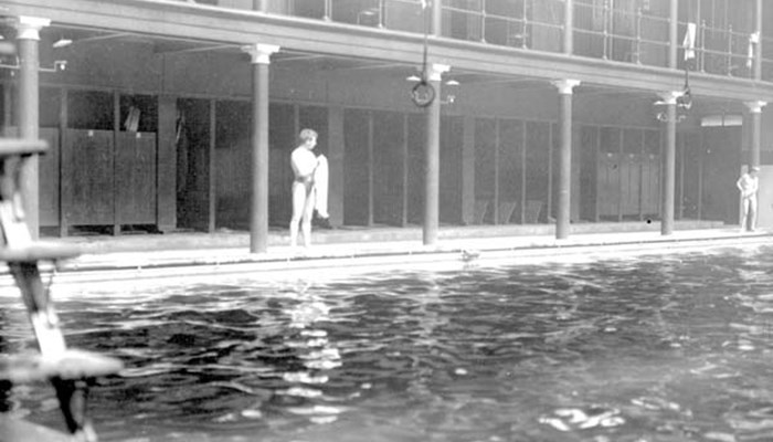 A black and white photo of the inside of a Glasgow swimming pool with the sun shining in from the windows and members about to get into the pool.