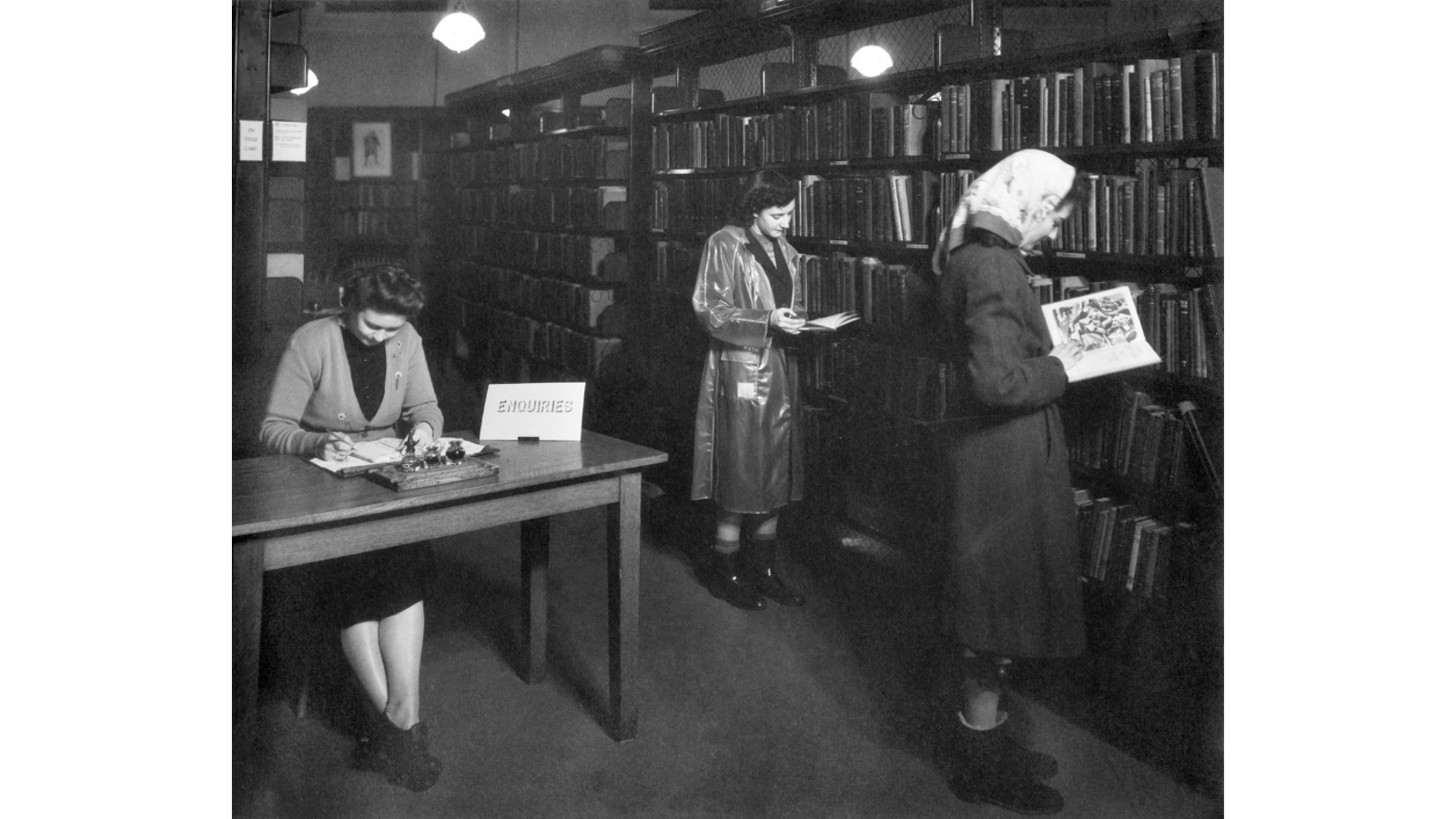 Black and white photograph of three women in a library: one writing at an enquiries desk while two others browse book stacks in the background.