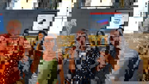 Four adults stand together indoors at a bar counter, smiling and holding up small tasting glasses.