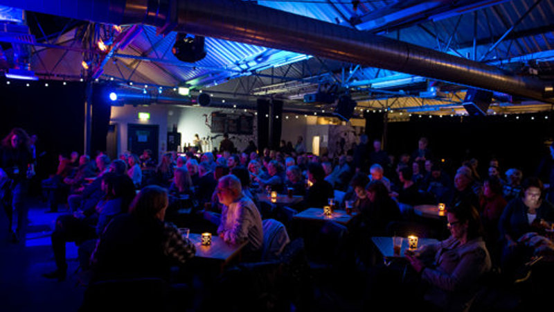 Audience seated at tables in a large industrial-style venue illuminated by blue stage lighting during a live event.