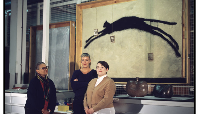 Three people stand in a museum store. Behind them is a large artwork showing the black silhouette of a leaping animal on a textured beige surface, framed in wood. In front of the artwork, two small sculptural ceramic objects with rounded, organic forms sit on a counter. The background includes metal mesh panels, drawers, pipes, and other industrial fixtures, giving the setting a functional, behind‑the‑scenes feel.