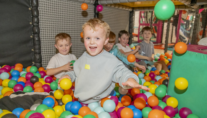 A young boy smiling while in a colourful soft play ball pit, four other young boys are behind them and there are balls flying everywhere.