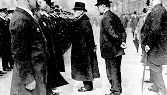 A large crowd of people well dressed in suits and uniforms including top hats with large buildings in the background.