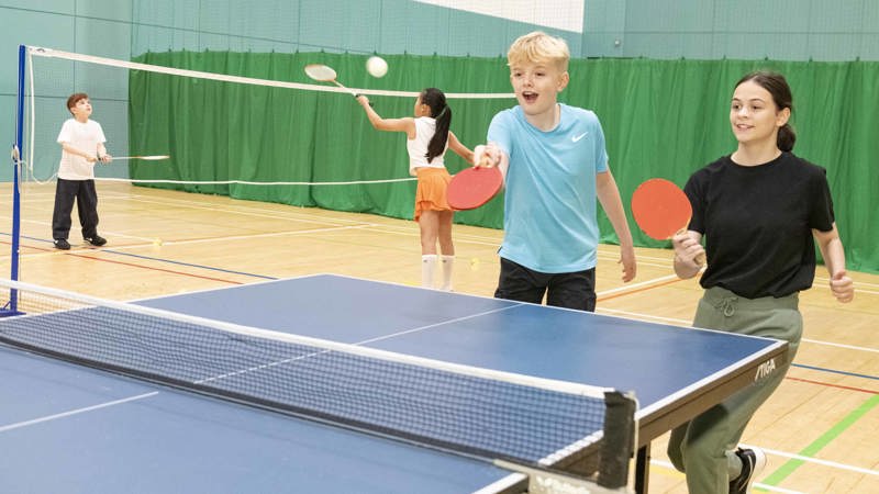 A boy and girl are playing tennis in an indoor court, in the background people are playing pickleball