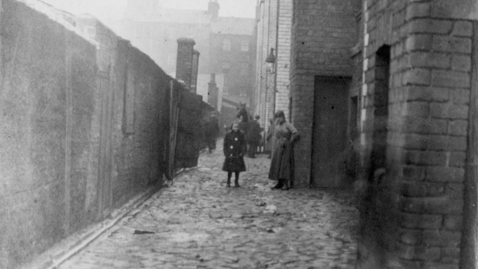 A black and white photograph of adults, children and horses in an alleyway between high brick buildings and a fence.