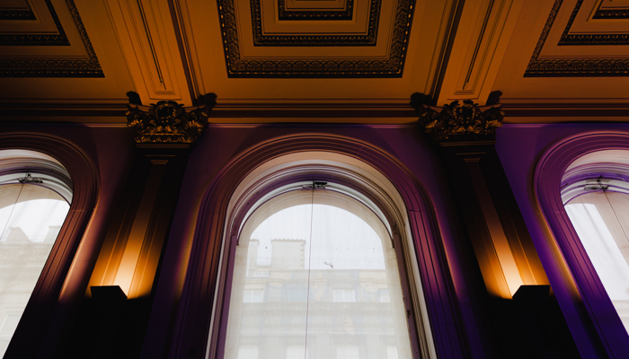 An ornate interior with three tall arched windows draped in sheer curtains, allowing a view of a building outside. Decorative columns, gold-accented moldings, and an intricately detailed ceiling frame the space, all illuminated by warm light that highlights the classical architectural details.