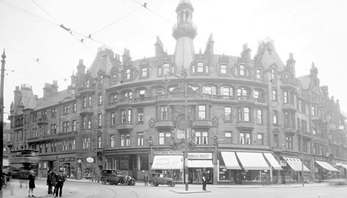 A black and white photo of a red sandstone building which is slightly curved round a corner of a cobbled road, tram lines are on the road and a tram is moving on the left hand side. Shop fronts are on the bottom row of the tall building.