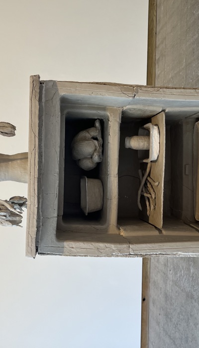 A monochromatic sculptural artwork made from a buff clay depicts a fridge without a door divided into compartments, each holding a different moulded object: a cooked chicken, a container, some beans and a juice carton. On top of the fridge is a vase with intricately sculpted drooping flowers and leaves. 