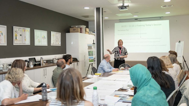 Glasgow Life Museums staff member presenting to a seated group in a meeting room with slides projected on a screen.