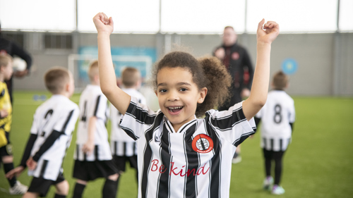 A child wearing a black and white football strip, smiles with arms raised above their head. Blurred out children in the same strip can be seen in the background.