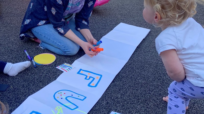 Artist Rebecca Fraser kneels over a home-made banner in a sunny playground, a toddler leaning over to talk to her