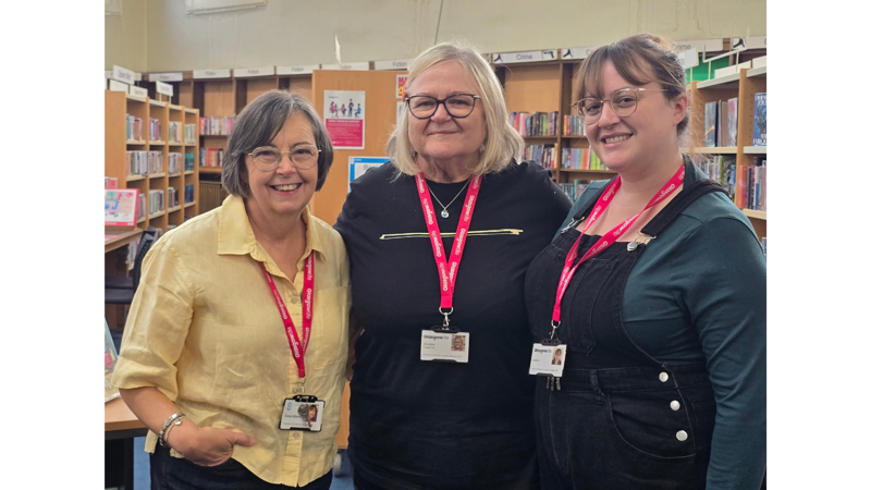 Three women posing together and smiling inside a library surrounded by bookshelves. The woman on the left wears a yellow button-up shirt and dark hair, the woman in the middle wears a black shirt and has blonde hair, and the woman on the right wears dark denim overalls over a teal long-sleeve shirt. All three are wearing glasses and red/pink lanyards with ID badges.