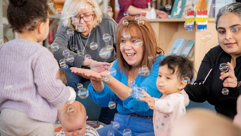 Group of adults and babies in a Glasgow Library setting, smiling and reaching out to catch bubbles which are floating in the air. There are bookcases and other people in the background.