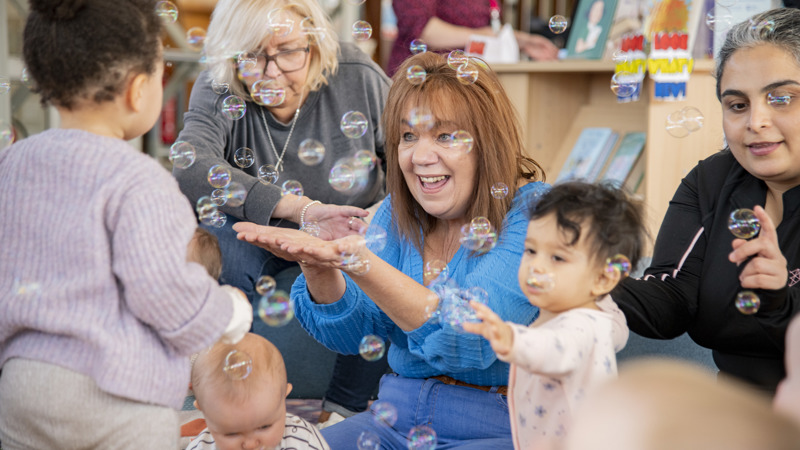 Group of adults and babies in a Glasgow Library setting, smiling and reaching out to catch bubbles which are floating in the air. There are bookcases and other people in the background.