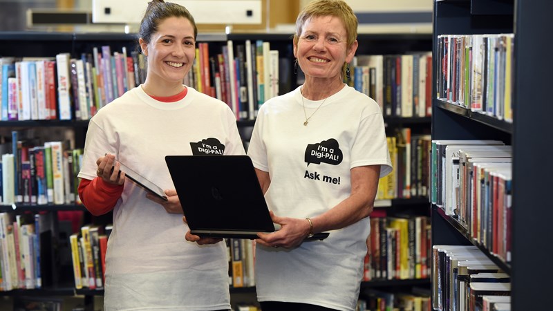 Two people in a library: one holding a laptop and one an ipad. Both are wearing white t-shirts that say 'I'm a digi pal - Ask me'