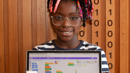 A young person wearing glasses and smiling holding up an open laptop in front of them showing a computer game