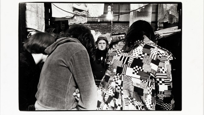 Black-and-white photo of a market stall with people browsing and patterned clothing in the foreground.