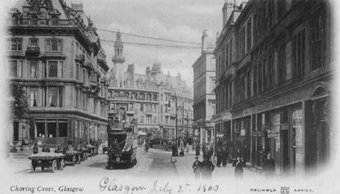A black and white postcard of a street in Glasgow with many carriages stocked with goods, people walking on the wide pavement and very tall buildings and a fountain.
