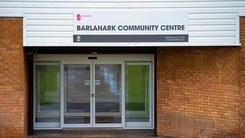 Exterior shot of Barlanark community centre. it is made of red brick and the sign is on a white background. the door is made up of glass with an additional pane of glass on each side of the door 