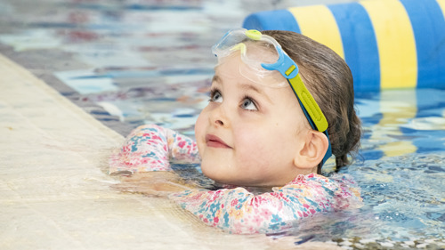 A young child looks upwards out of a swimming pool with goggles on their head.