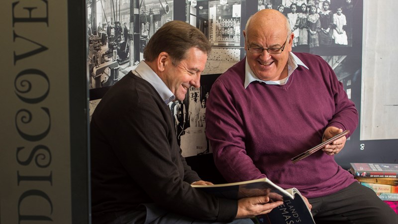 two men sitting next to each other looking at a book and smiling