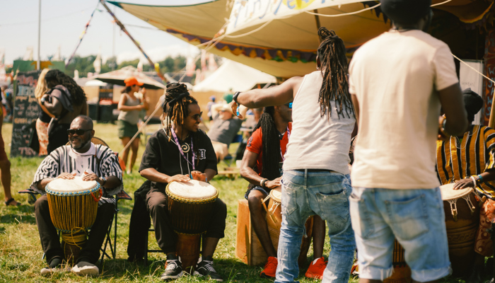 A group of musicians sit in a row playing djembe drums on a grassy area near a "Tiny Tea Tent" while onlookers watch.