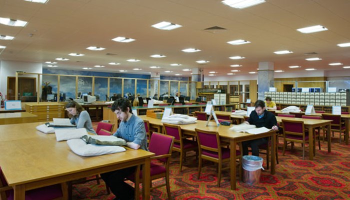 A photograph of people sitting in the search room on large pine tables and red upholstered chairs looking at large paperworks.