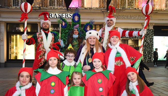 Performers dressed in red, white and green festive clothing and costumes stand together in front of Christmas lights
