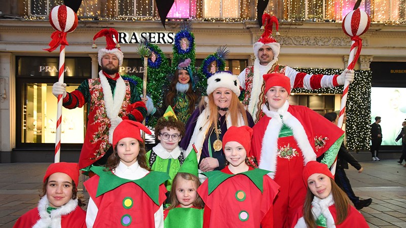 Performers dressed in red, white and green festive clothing and costumes stand together in front of Christmas lights