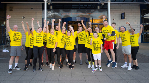 A large group of people gathered for a photo outside a leisure centre. They are all wearing the same yellow t-shirts and have their fists raised in celebration