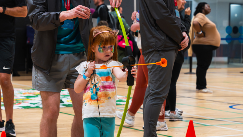 A member of staff helps a young girl with red hair and her face painted as a butterfly with a bow and arrow during archery at a family fun day.