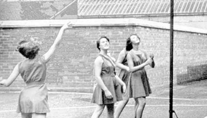 A black and white photo of a small group of people playing netball outside. One person has took a shot and the ball is in the air going towards the hoop.