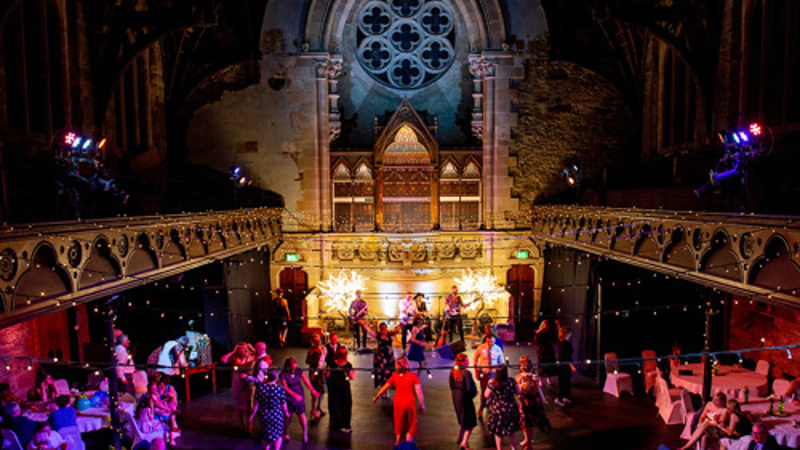 Guests dancing and socialising in a grand church hall transformed for an evening event, with colourful lighting and a stage backdrop.