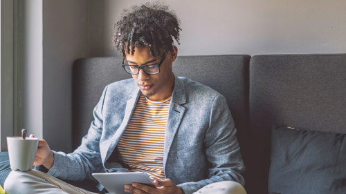 A person wearing glasses sitting crossed legged on a sofa. They are reading from an ipad and holding a mug of coffee.
