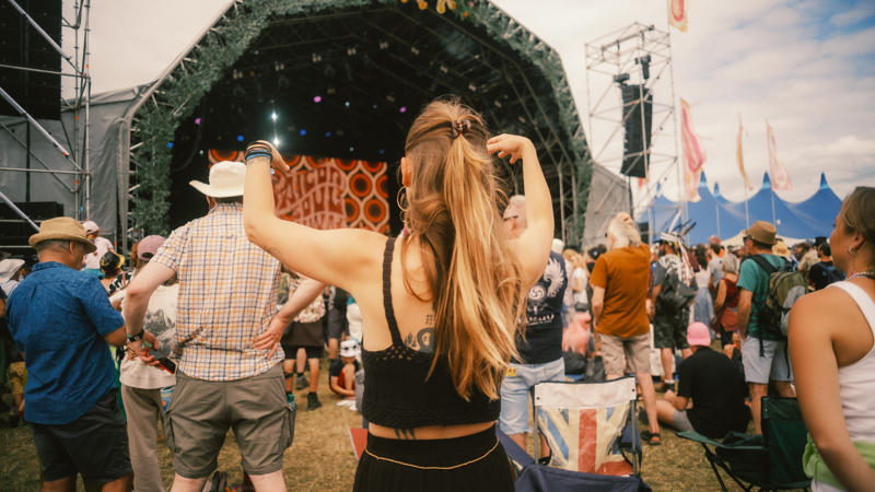 A person with their hair in a ponytail stands with their back to the camera, looking toward a large WOMAD festival stage decorated with greenery.
