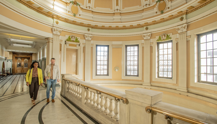 A women and a man walking along a corridor in the Mitchell Library with light shining through the roof